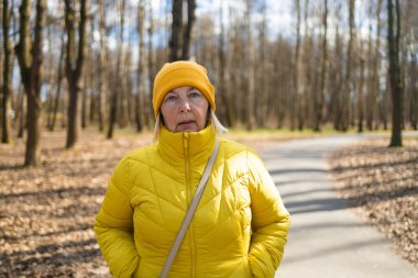 Sarı ceket ve şapka giymiş düşünceli bir kadın, sonbahar ağaçları ve yapraklarla çevrili bir park yolunda duruyor. Huzurlu sahne, değişen mevsimlerin güzelliğini yakalar.