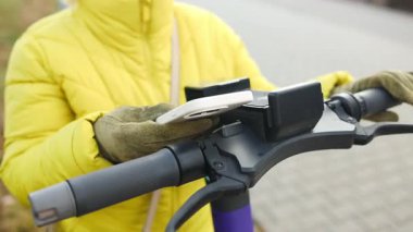 Sarı ceketli ve bereli bir kadın şehir parkında elektrikli scooter kiraladı. Olay yeri, çevre dostu taşımacılık için uygulama bazlı kiralama hizmetlerinin kolaylığını yakalıyor.