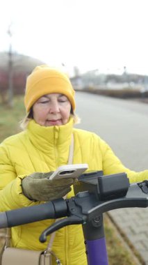 Sarı ceketli ve eldivenli bir kadın elektrikli scooterının yanında dururken telefonunu tutuyor. Görüntü, mobilet ve telefon odaklı modern, aktif yaşam tarzını yakalar.