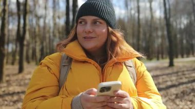 Sarı ceketli ve siyah bereli mutlu bir kadın parkta elinde telefonuyla yürüyor. Parlak doğal ortam, ağaçlar ve huzurlu bir yol, o anın neşeli havasını arttırır..