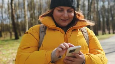 Sarı ceketli ve siyah bereli mutlu bir kadın parkta elinde telefonuyla yürüyor. Parlak doğal ortam, ağaçlar ve huzurlu bir yol, o anın neşeli havasını arttırır..