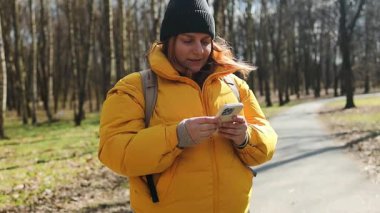 Sarı ceketli ve siyah bereli mutlu bir kadın parkta elinde telefonuyla yürüyor. Parlak doğal ortam, ağaçlar ve huzurlu bir yol, o anın neşeli havasını arttırır..