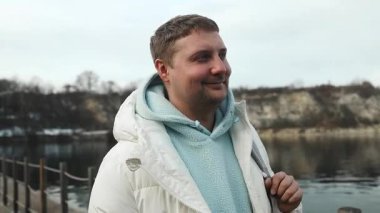 Dağlarda nehir kıyısında kameraya bakarak tek başına duran gülümseyen yakışıklı beyaz adamın portresi. Genç erkek kameraya bakıyor ve gülümsüyor.. . 