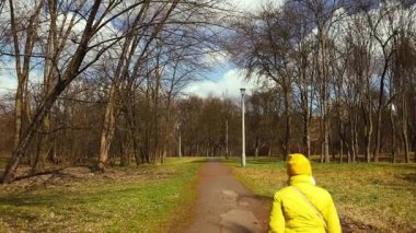 Sarı ceketli bir adam elinde mavi bir çantayla parkta yürür. Görüntü, parkın canlı renklerini ve değişen mevsimleri gösteren, doğada barışçıl bir yürüyüş görüntüsü yakalıyor.