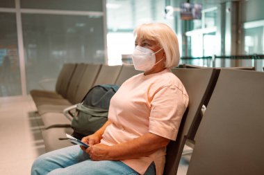 Tıbbi maske takan yaşlı bir kadın havaalanı bekleme odasında oturuyor ve sosyal mesafeyi gözetliyor. Yüksek kalite fotoğraf