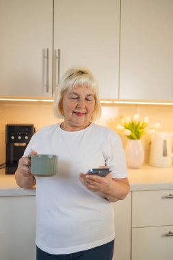 Son sınıf öğrencisi bir kadın elinde telefon ve kahve fincanıyla mutfakta durup gülümsüyor. Bu resim, yaşlıların modern yaşamlarının teknoloji ve rahatlıkla günlük rutinlerinin tadını çıkardıklarını vurguluyor.