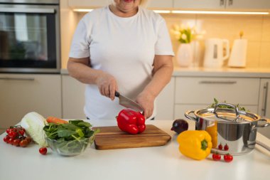 Bir kadın mutfaktaki tahta kesme tahtasında taze kırmızı bir dolma biber doğruyor. Etrafında havuç, yeşil yapraklar ve taze sebzelerle dolu bir tencere var.