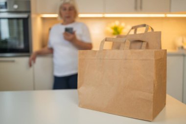 Son sınıf öğrencisi bir kadın mutfakta durup telefonuna bakıyor. Önünde masanın üstünde iki tane poşet var, muhtemelen teslim edilmiş ya da alınmış, açılmaya hazır..