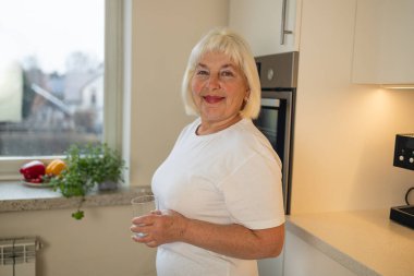 Mutfakta elinde bir bardak suyla gülümseyen neşeli bir son sınıf öğrencisi. Bu görüntü sağlıklı yaşamanın özünü ve evde susuz kalmanın zevkini yansıtıyor.