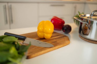Taze sarı bir dolma biber mutfaktaki ahşap bir kesme tahtasına oturur. Yanında bir bıçak, diğer sebzeler ve arka planda bir tencere var..