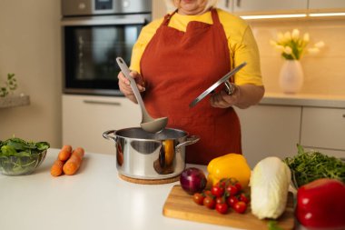 Sarı önlüklü yaşlı bir kadın mutfak tezgahında taze havuç, domates ve diğer malzemelerle sebzeleri karıştırıyor..