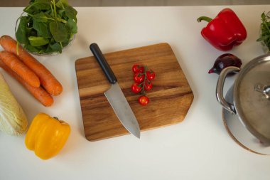 Taze vişneli domatesler parlak bir mutfakta ahşap bir kesme tahtasının üzerinde dilimlenir. Keskin bir bıçak kullanılır ve havuç, dolma biber ve ıspanak gibi renkli sebzeler yakınlardadır..