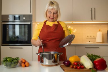 Sarı önlüklü yaşlı bir kadın mutfak tezgahında taze havuç, domates ve diğer malzemelerle sebzeleri karıştırıyor..