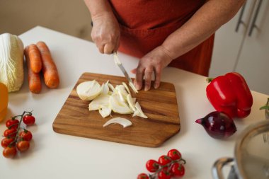 Mutfakta tahta kesme tahtasıyla soğan doğrayan biri. Yakınlarda, domates, havuç ve biber gibi çeşitli taze sebzeler yayılarak pişirmeye hazırlanıyor.