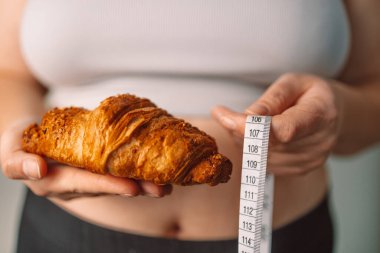Bir elinde kruvasan, diğer elinde mezura tutan bir kadın, hoşgörülü yiyeceklerin sağlık ve sağlık hedefleri ile dengelenmesinin zorluğunu yansıtıyor. Diyet yapma ve kendini kontrol etme kavramı
