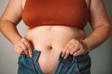 Dar kotları iliklemeye çalışan bir kadın vücut görüntüsü ve kilo sorunlarını sembolize ediyor. Bu, çoğu kişi arasında yaygın olarak görülen kişisel imajın ve kıyafetlere uymanın günlük mücadelesini yansıtır.