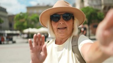 60 'ların son sınıf öğrencisi şapkalı ve güneş gözlüklü bir kadın canlı bir şehir merkezinde selfie çekiyor. O, güneşli bir günde popüler bir turistik yeri ziyaret ederken seyahat anlarının tadını çıkarıyor..