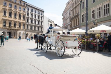 Krakow, Polonya - Temmuz, 2024: Beyaz at arabası arka planda tarihi binalar bulunan hareketli bir şehir meydanında hareket ediyor. Turistler ve yerliler etrafta dolanıyor ve açık hava kafeleri...
