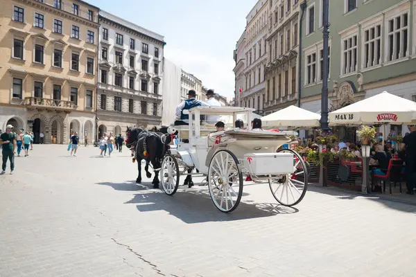 Krakow, Polonya - Temmuz, 2024: Beyaz at arabası arka planda tarihi binalar bulunan hareketli bir şehir meydanında hareket ediyor. Turistler ve yerliler etrafta dolanıyor ve açık hava kafeleri...