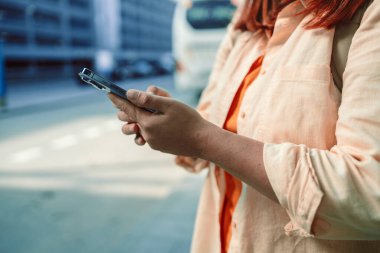 Kızıl saçlı bir kadın dışarıdaki ekrana bakarken akıllı telefonunu elinde tutuyor. Bu resim, kentsel etkileşim, mesajlaşma veya navigasyon için akıllı telefon kullanımını vurgular