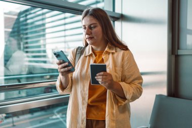 Havaalanı terminalinde elinde pasaportu olan genç bir kadın akıllı telefonuna bakıyor ve uçuşuna hazırlanıyor. Modern havaalanı içerisi onun arkasındaki pencereden görülebiliyor..