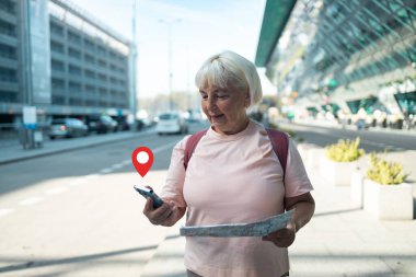 Yaşlı bir kadın elinde bir haritayla havaalanında geziniyor ve bir sonraki hedefini arıyor. GPS konum pin simgesi onun dijital navigasyon kullanımını vurgulamak için resmin üzerine yerleştirilir