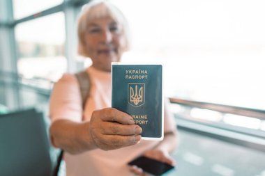 Kıdemli kadın havaalanı terminalinde duruyor, gülümsüyor ve elinde pasaportu ve akıllı telefonu var. Modern havaalanı pencereleri ve parlak, hoş bir atmosferi olan uçuşu için hazır.