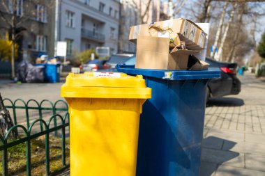 İki çöp kutusu, biri sarı ve biri mavi, kaldırıma yerleştirilmiş. Kutular, arka planda yeşillik ve kentsel binalarla çevrili karton kutular ve atıklarla dolu. Temiz bir cadde ve