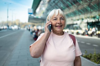 Son sınıf öğrencisi kadın havaalanı terminalinin önünde gülümseyerek telefonla konuşuyor. Parlak güneş ışığı seyahat ederken onun neşesini vurguluyor. Günlük kıyafetleri ile uyumlu.