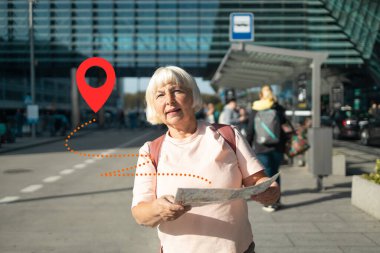 Yaşlı kayıp bir kadın elinde bir haritayla havaalanında geziniyor ve bir sonraki hedefini arıyor. GPS konum pin simgesi onun dijital navigasyon kullanımını vurgulamak için resmin üzerine yerleştirilir