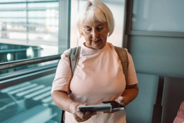 Son sınıf öğrencisi bir kadın havaalanı terminalinde akıllı telefonunu tutarken gülümsüyor. Uçuşunu beklerken, terminalin büyük cam pencerelerinin önünde duruyor. Modern havaalanı içi görünür..