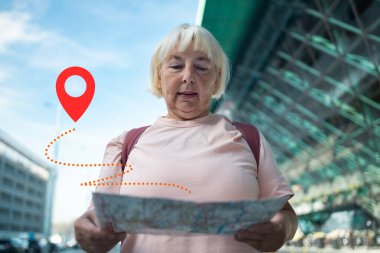 Yaşlı bir kadın elinde bir haritayla havaalanında geziniyor ve bir sonraki hedefini arıyor. GPS konum pin simgesi onun dijital navigasyon kullanımını vurgulamak için resmin üzerine yerleştirilir