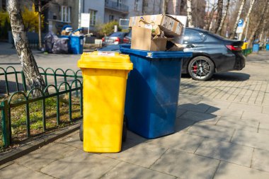 İki çöp kutusu, biri sarı ve biri mavi, kaldırıma yerleştirilmiş. Kutular, arka planda yeşillik ve kentsel binalarla çevrili karton kutular ve atıklarla dolu. Temiz bir cadde ve