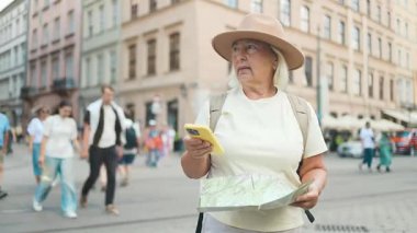 Bir şehir caddesinde elinde harita tutan şapkalı yaşlı bir kadın yerel simgeleri keşfediyor, tarihi merkezde seyahat ediyor. Parlak güneşli yaz günü, tam şehir manzarası.