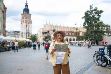 Sarı ceketli ve şapkalı bir kadın canlı bir şehir meydanında elinde haritayla bölgeyi dolaşmaya çalışıyor. Şehir mimarisi ve bisikletçileriyle turist gibi görünüyor.