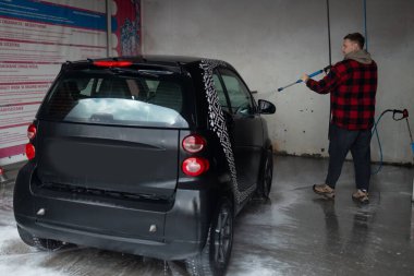 Sakallı bir adam oto yıkamada yüksek basınçlı çamaşır makinesiyle gri bir arabayı yıkar. Pahalı reklam fotoğrafçılığı