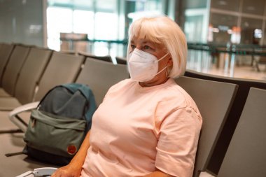 Tıbbi maske takan yaşlı bir kadın havaalanı bekleme odasında oturuyor ve sosyal mesafeyi gözetliyor. Yüksek kalite fotoğraf