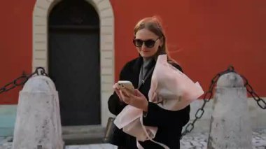 Uzun saçlı, güneş gözlüğü takan genç bir kadın elinde bir buket çiçekle dışarıda telefonunu kontrol ediyor. Arka planda canlı turuncu bir duvar var. Kapısı ve dekoratif taşları var.