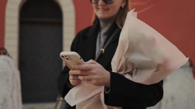 Uzun saçlı, güneş gözlüğü takan genç bir kadın elinde bir buket çiçekle dışarıda telefonunu kontrol ediyor. Arka planda canlı turuncu bir duvar var. Kapısı ve dekoratif taşları var.