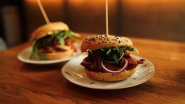 Beyaz tabakta iki lezzetli gurme burger, üzerinde çıtır pastırma, taze yeşillikler ve ahşap bir masaya konmuş soğanlar. Yumuşak susamlı çörekler kolay dokunmak için bir kürdanla şişlenir. Şey...