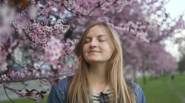 Gülümseyen bir kadın baharda çiçek açan kiraz çiçeklerinin önünde duruyor. Canlı pembe çiçekler, güzel bir açık hava sezonunun tadını çıkarırken huzurlu ve resimsel bir atmosfer yaratıyor.