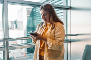 Sarı elbiseli genç bir kadın havaalanı terminalinde uçuşunu beklerken akıllı telefonunu kontrol ediyor. Havaalanlarında yaygın bir faaliyet olan modern teknolojiyi kullanmakla meşgul.