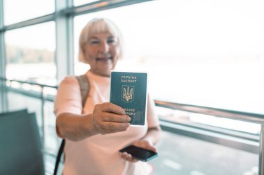 Kıdemli kadın havaalanı terminalinde duruyor, gülümsüyor ve elinde pasaportu ve akıllı telefonu var. Modern havaalanı pencereleri ve parlak, hoş bir atmosferi olan uçuşu için hazır.