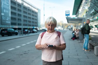 Kızıl saçlı bir kadın dışarıdaki ekrana bakarken akıllı telefonunu elinde tutuyor. Bu resim, kentsel etkileşim, mesajlaşma veya navigasyon için akıllı telefon kullanımını vurgular