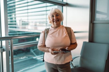 Son sınıf öğrencisi bir kadın havaalanı terminalinde akıllı telefonunu tutarken gülümsüyor. Uçuşunu beklerken, terminalin büyük cam pencerelerinin önünde duruyor. Modern havaalanı içi görünür..