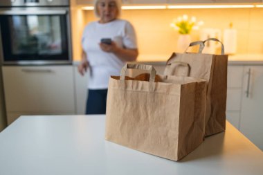 Son sınıf öğrencisi bir kadın mutfakta durup telefonuna bakıyor. Önünde masanın üstünde iki tane poşet var, muhtemelen teslim edilmiş ya da alınmış, açılmaya hazır..