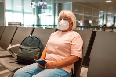 Tıbbi maske takan yaşlı bir kadın havaalanı bekleme odasında oturuyor ve sosyal mesafeyi gözetliyor. Yüksek kalite fotoğraf