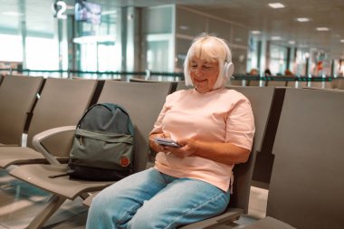 Havaalanı terminalinde tatil için uçağa binmeyi bekleyen 50 'li yaşlarda beyaz çekici bir kadın. Akıllı telefon kullanıyor, müzik dinlemek için kulaklık takıyor yanında bavul taşıyor.
