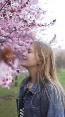 Gülümseyen bir kadın baharda çiçek açan kiraz çiçeklerinin önünde duruyor. Canlı pembe çiçekler, güzel bir açık hava sezonunun tadını çıkarırken huzurlu ve resimsel bir atmosfer yaratıyor.