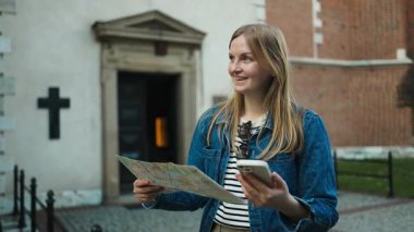 Polonya 'nın Krakow kentindeki tarihi yerleri keşfetmek için bir harita ve akıllı telefon kullanarak gülümseyen kadın gezgin. Kilisenin girişinde duruyor, eski kasabada bahar gününün tadını çıkarıyor..